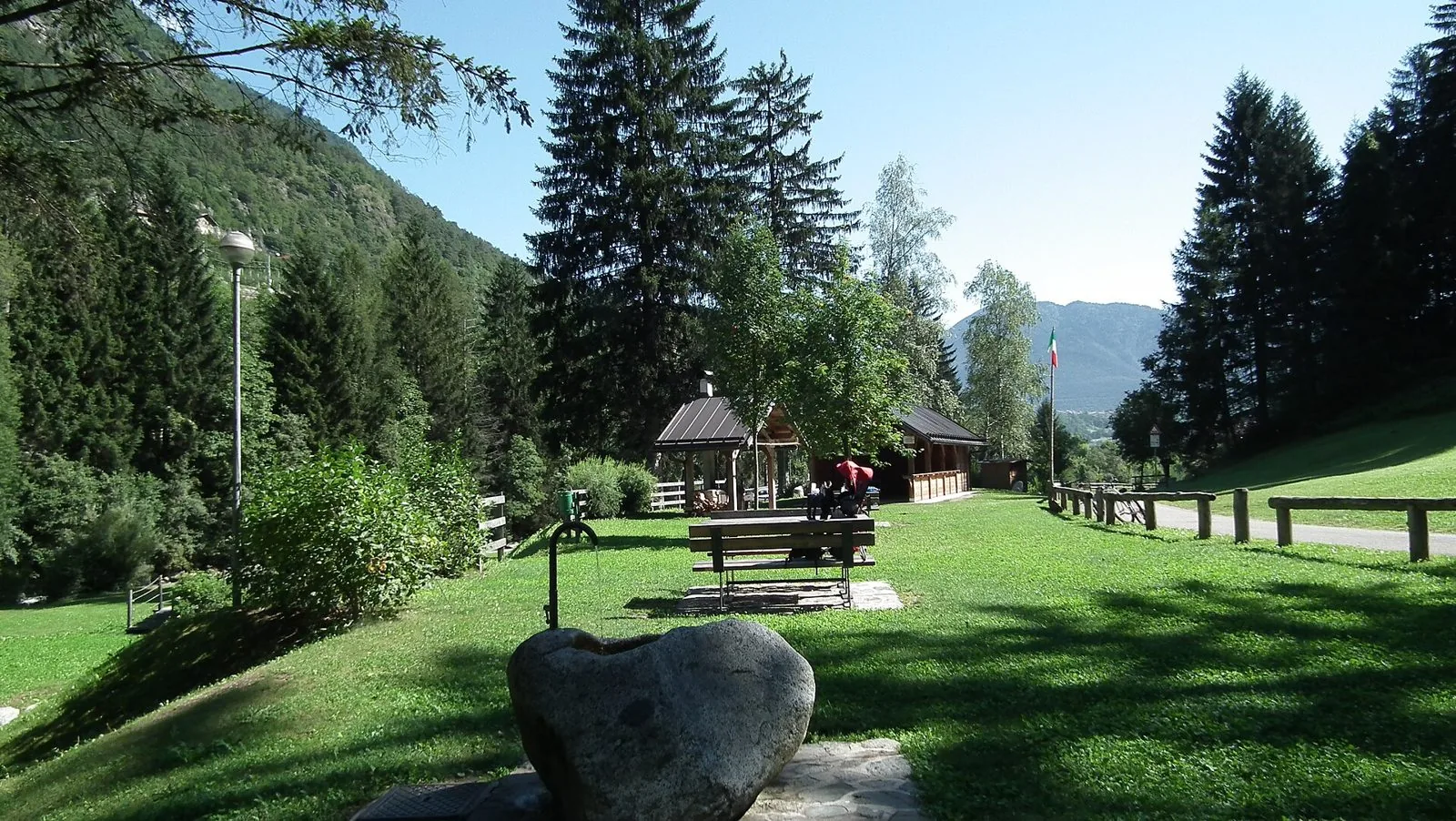 Un'area di sosta con prato, panchine e fontana lungo la pista ciclabile della Val di Sole, tra abeti e montagne