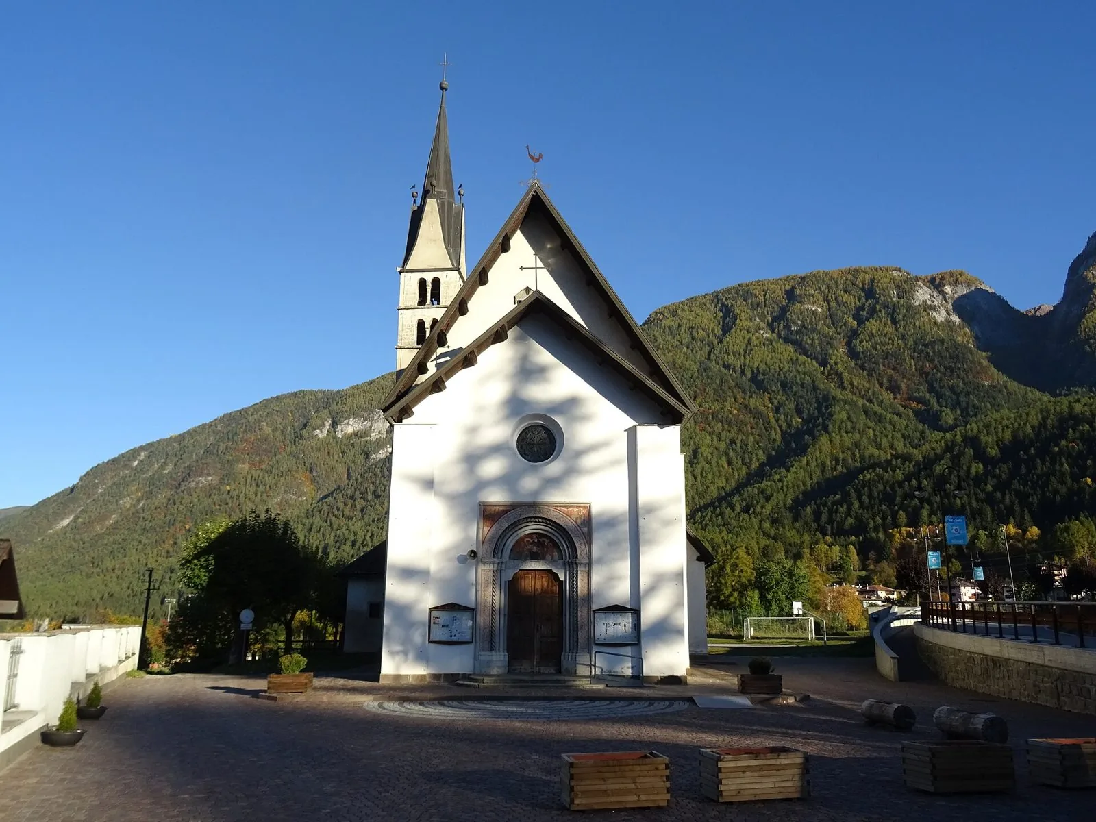 La facciata bianca della chiesa di San Lorenzo a Dimaro con il campanile a cuspide e i boschi della Val di Sole sullo sfondo