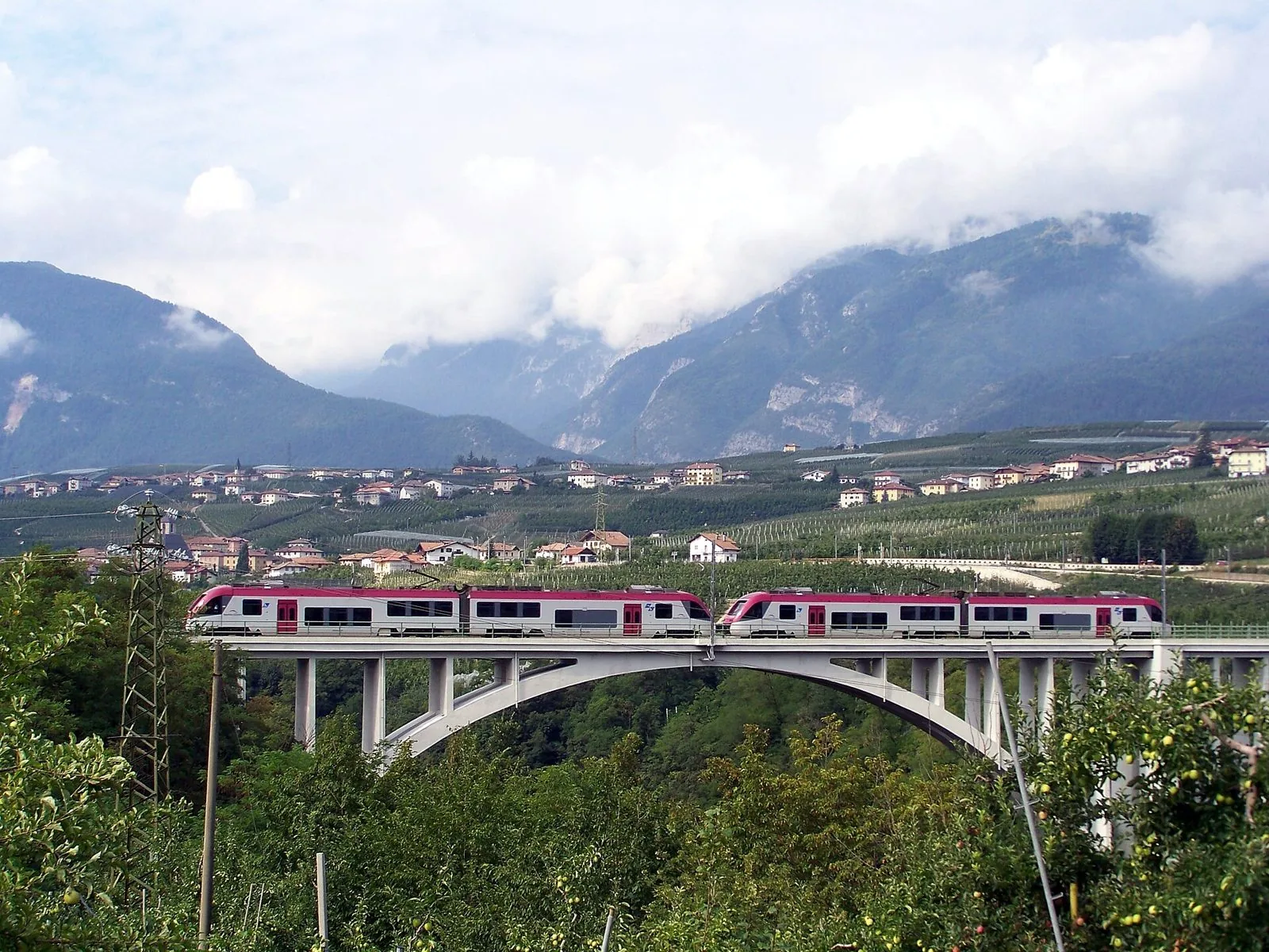 Due convogli elettrici bianchi e rossi della ferrovia Trento-Malè sul ponte ad arco di Santa Giustina, tra frutteti e montagne